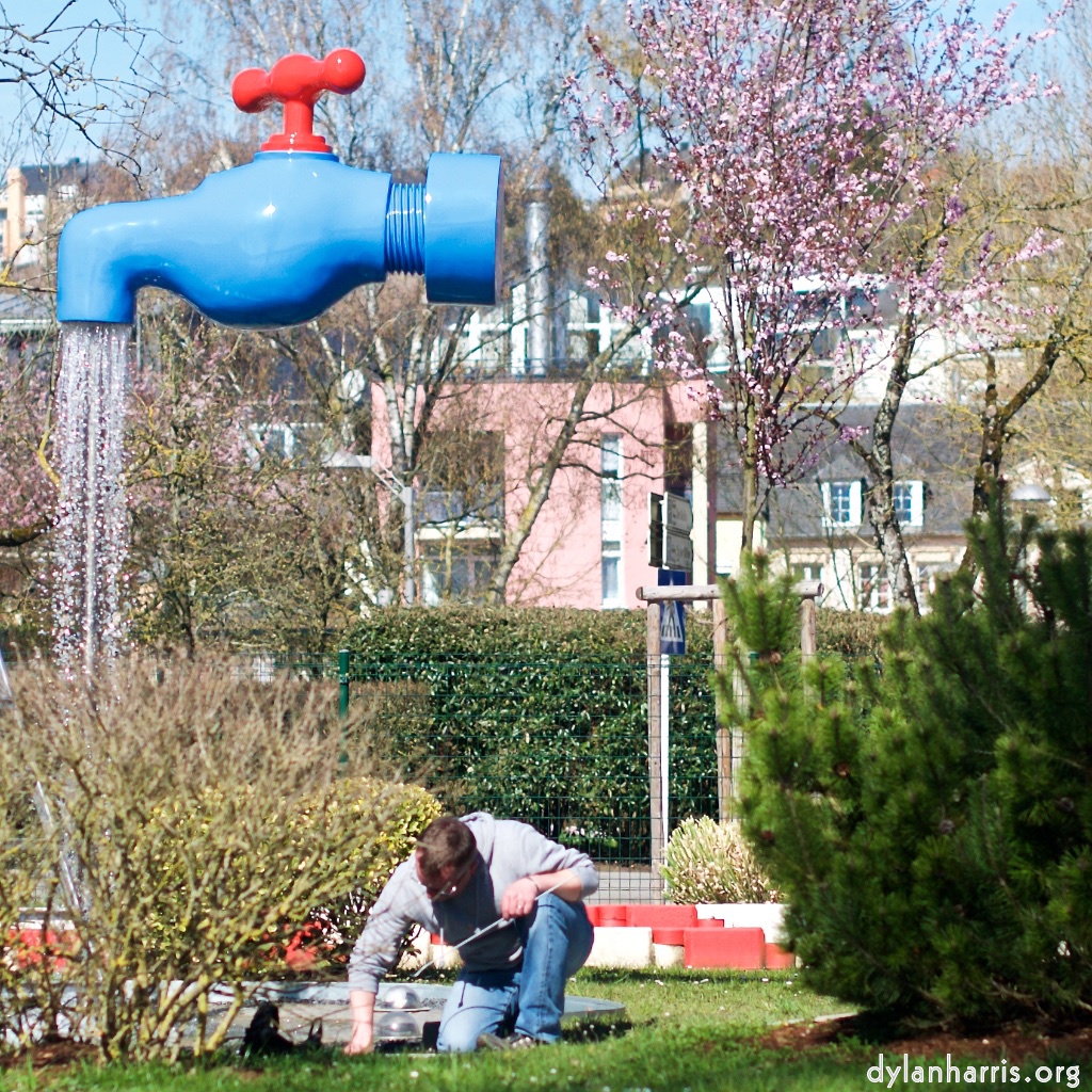 image: a tap, floating in the air, watering a small shrub, whilst a bloke gardens.